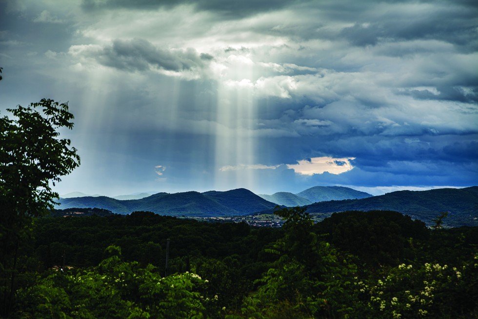 Cypress and olive trees at the town’s edge, and a break in the clouds over mountains.